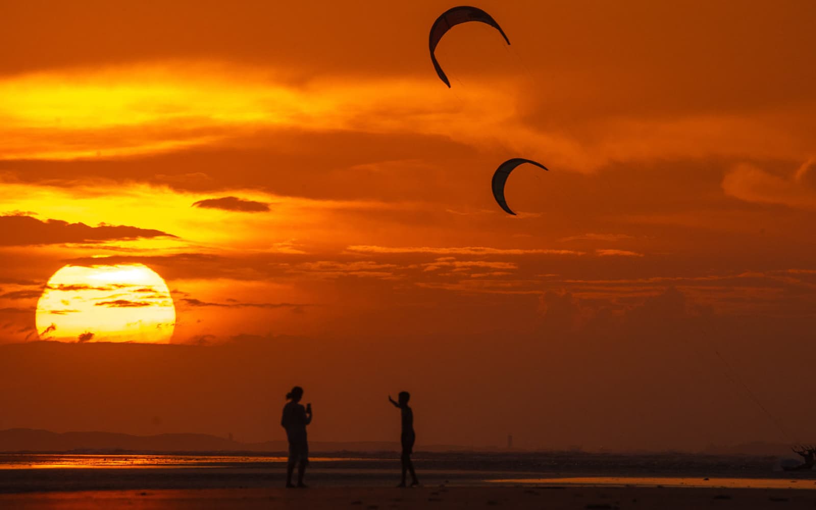 Kites no céu ao pôr do sol, com silhuetas na praia