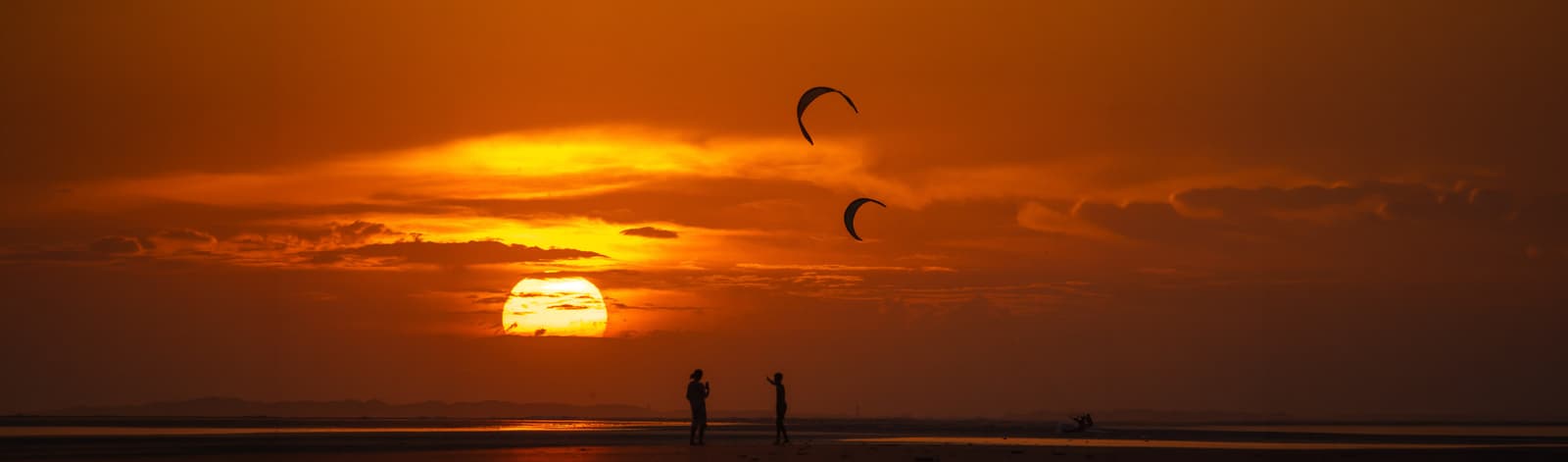 Kites no céu ao pôr do sol, com silhuetas na praia